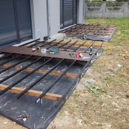 SHINE WOOD - Budowa tarasu: konstrukcja z legarów stalowych i drewnianych, częściowo pokryta deskami, widoczne narzędzia i materiały budowlane, czarna geowłóknina, dom w tle.