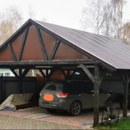 Zadaszone miejsce parkingowe z drewnianą konstrukcją i brązowym dachem krytym blachą falistą, pod spodem zaparkowany samoch&oacute;d osobowy, widoczne narzędzia i sprzęt ogrodniczy.