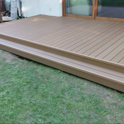 SHINE WOOD - Beżowy taras z desek kompozytowych z dwoma stopniami, przylegający do budynku z oknem tarasowym, otoczony trawnikiem. Widok z niskiej perspektywy.