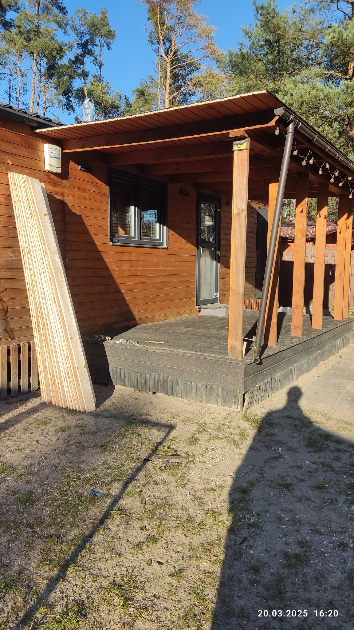 Ciemny taras przy drewnianym domku z bali, widoczne deski przygotowane do montażu, cień osoby fotografującej na pierwszym planie.