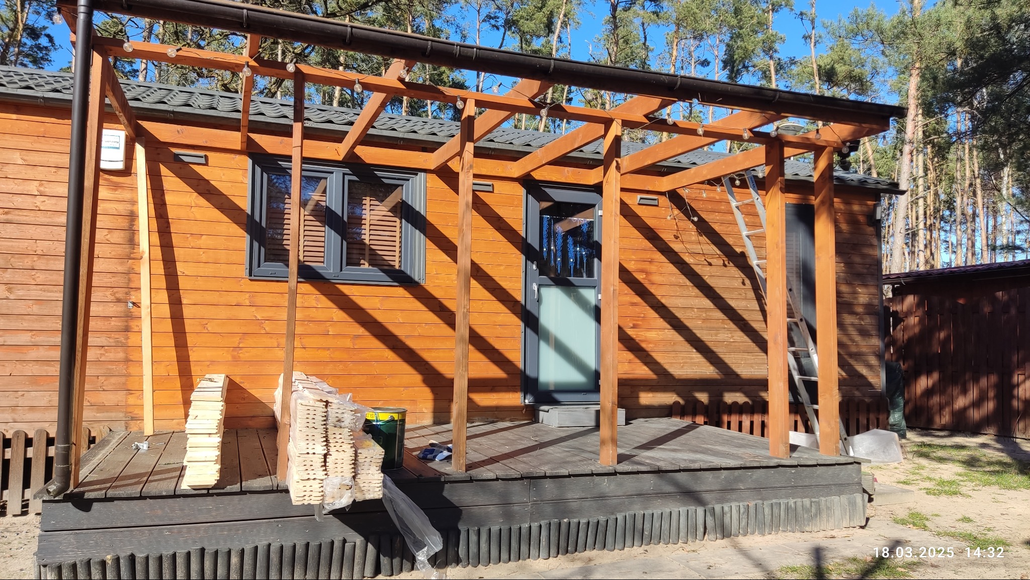 Drewniana konstrukcja zadaszenia nad tarasem w trakcie budowy, widoczne deski, drabina i narzędzia, tło stanowią drzewa.