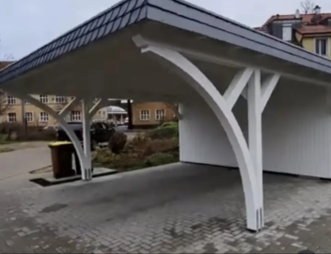 Drewniana wiata garażowa z białymi, łukowatymi podporami i ciemnoszarym dachem z dachówki bitumicznej, widoczna z zewnątrz na tle zabudowań mieszkalnych.