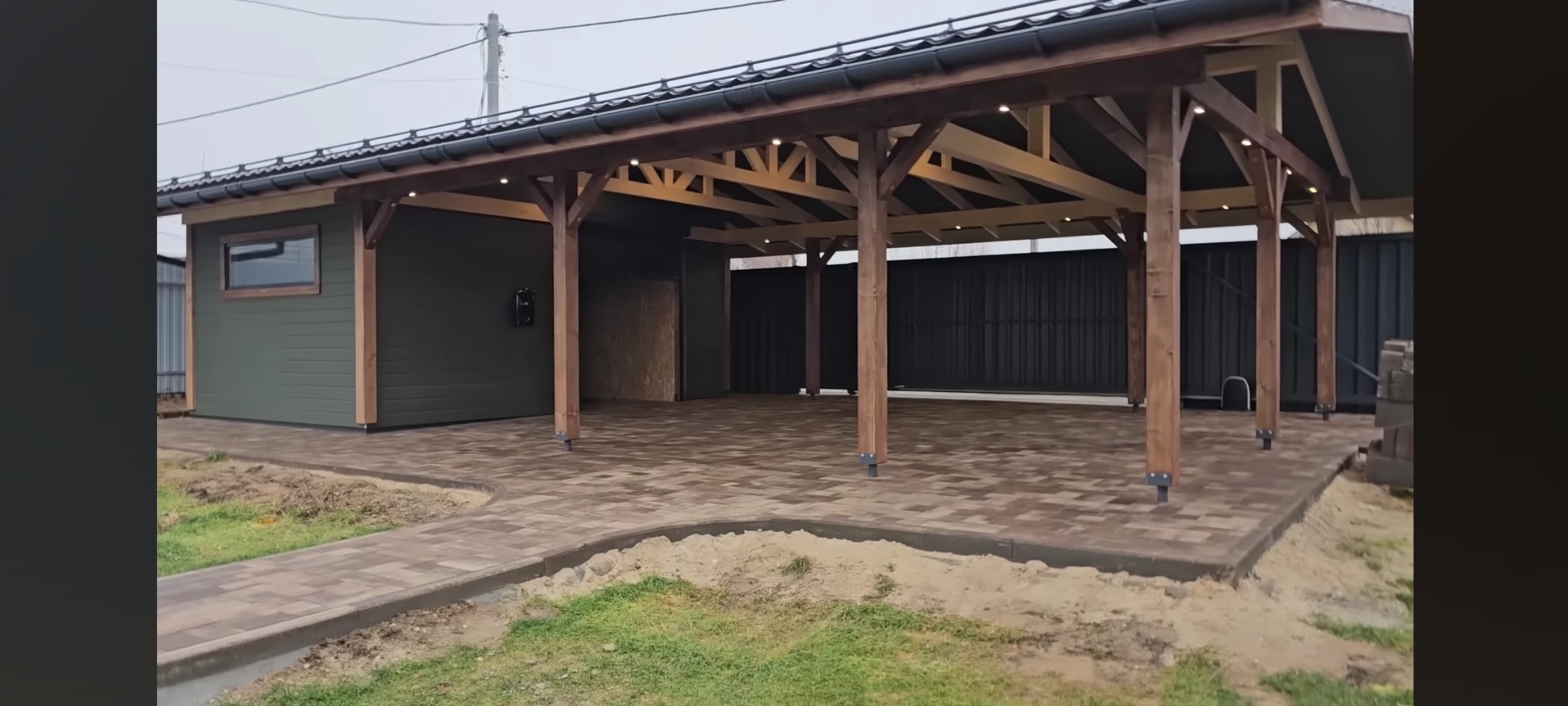 Zadaszona konstrukcja z drewnianymi słupami i oświetleniem punktowym, przylegająca do budynku z zieloną elewacją, wyłożona kostką brukową, w tle metalowy płot.