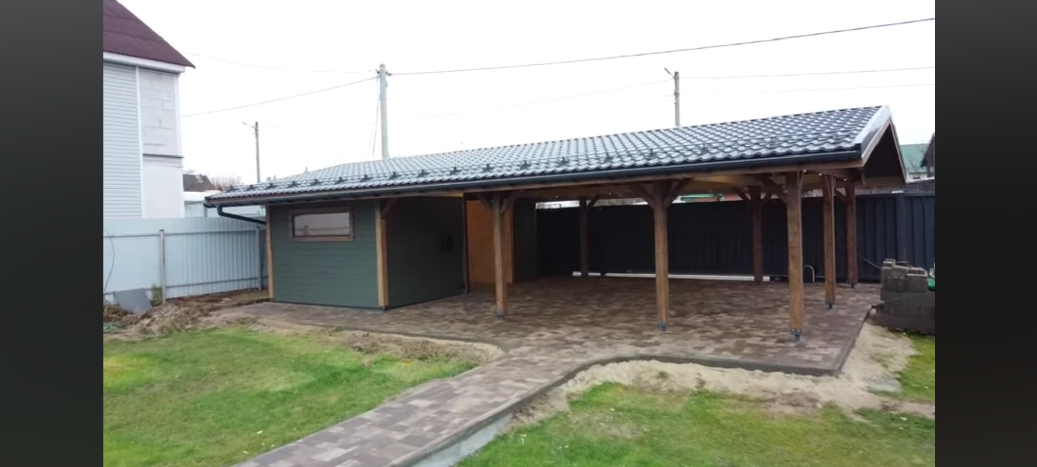 Zadaszona konstrukcja z drewnianymi słupami i zamykanym pomieszczeniem obok, wyłożona kostką brukową, z ścieżką z kostki prowadzącą od trawnika.