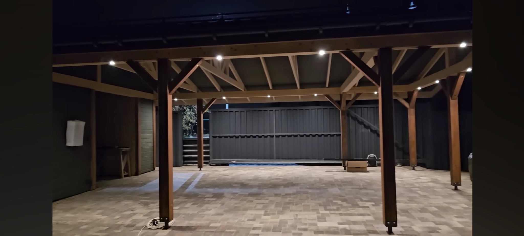 Zadaszona konstrukcja z drewnianych belek z oświetleniem punktowym, wyłożona kostką brukową, z widoczną ścianą i fragmentem ogrodzenia w tle.