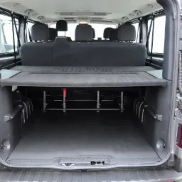 Wnętrze szarego busa z otwartymi tylnymi drzwiami, widoczny rząd siedzeń i przestrzeń bagażowa z metalową konstrukcją pod podwyższoną półką.