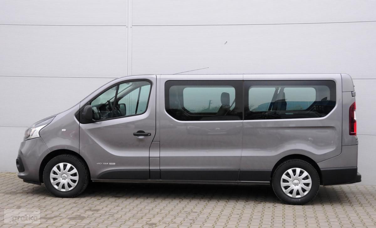 Szary, ośmioosobowy bus Renault Trafic z przyciemnianymi szybami, zaparkowany na kostce brukowej przed szarą ścianą hali.