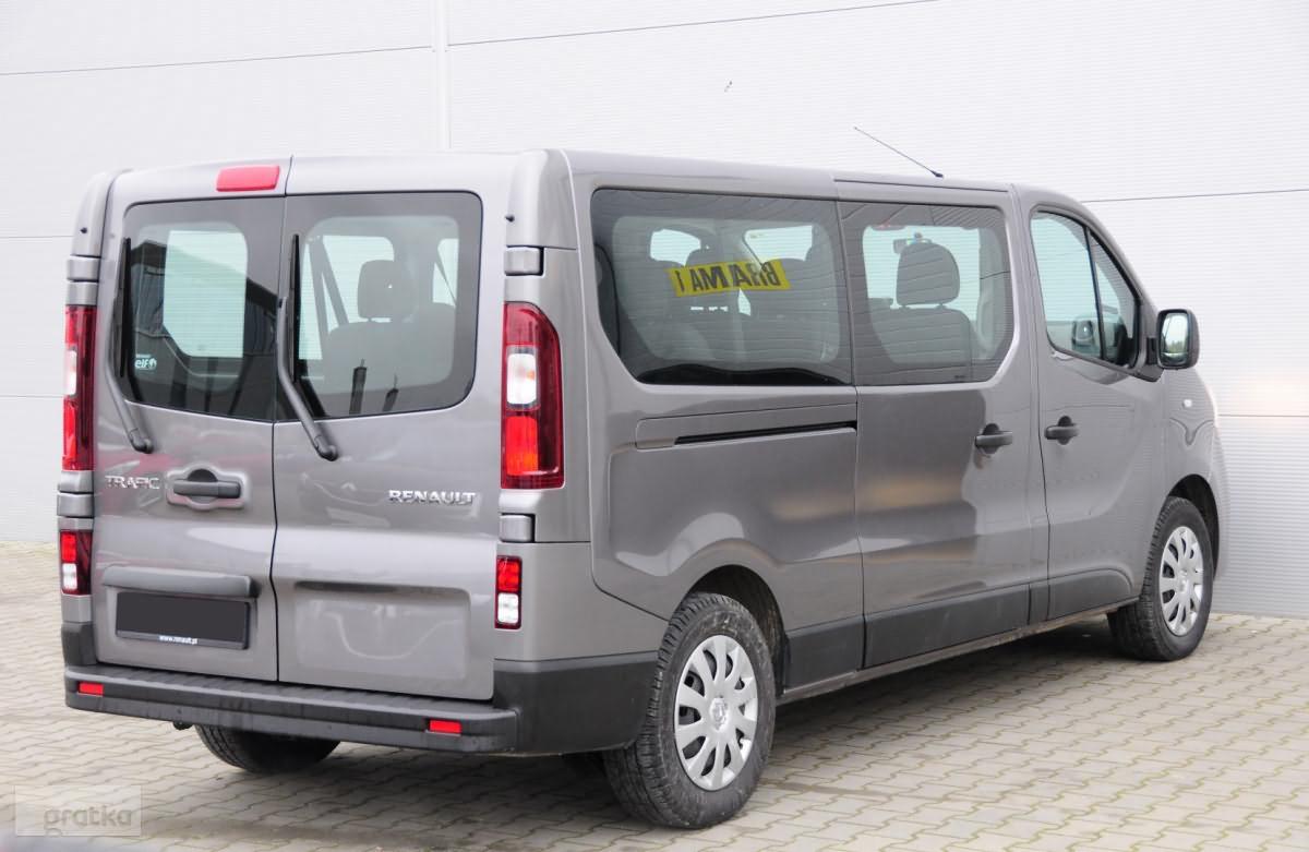 Szary samochód dostawczy Renault Trafic z widoczną naklejką na szybie, zaparkowany na kostce brukowej przed jasną ścianą.