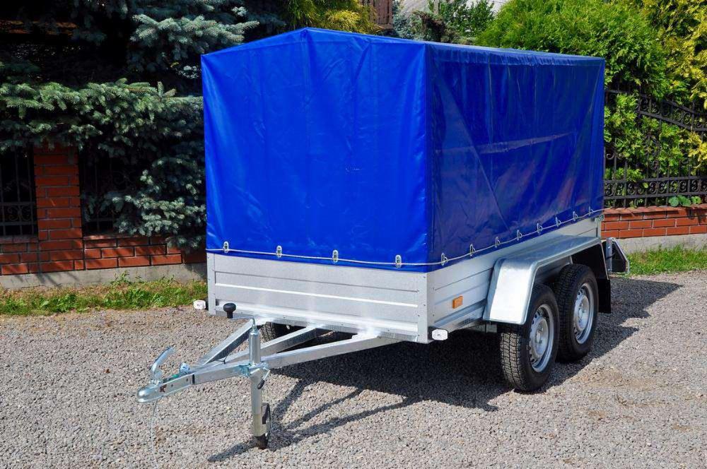 Dwukołowa przyczepa transportowa z niebieską plandeką, zaparkowana na żwirowym podłożu, widoczna na tle ceglanego muru i zielonych krzewów.