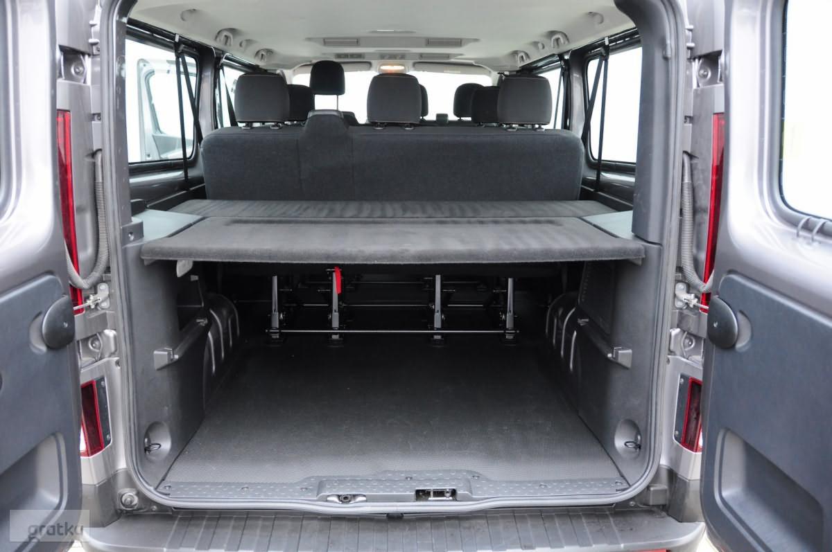 Wnętrze szarego busa z otwartymi tylnymi drzwiami, widoczny rząd siedzeń i przestrzeń bagażowa z metalową konstrukcją pod podwyższoną półką.