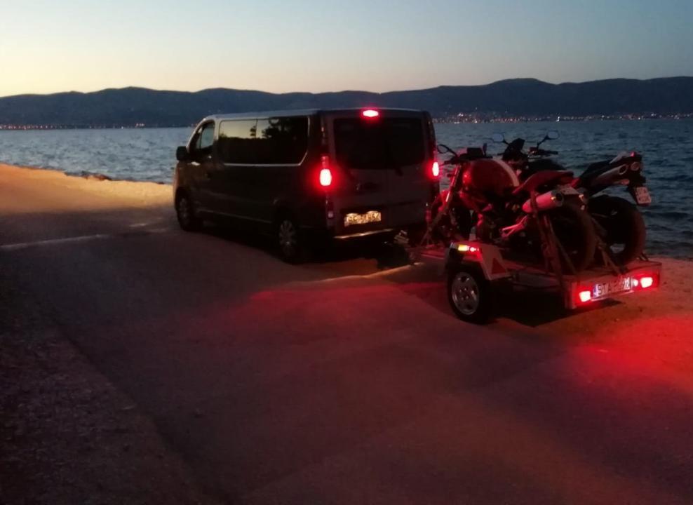 Czarny van z przyczepą przewożącą dwa motocykle, zaparkowany na poboczu drogi nad brzegiem morza o zmierzchu, z widokiem na góry w oddali.