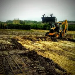 Żółta koparko-ładowarka z podniesioną łyżką na rozjeżdżonym, błotnistym terenie budowy, w tle las i pochmurne niebo.