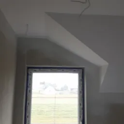 Pokój na poddaszu po nałożeniu gładzi, widoczne kable wystające ze ściany i sufitu, okno z widokiem na zewnątrz.