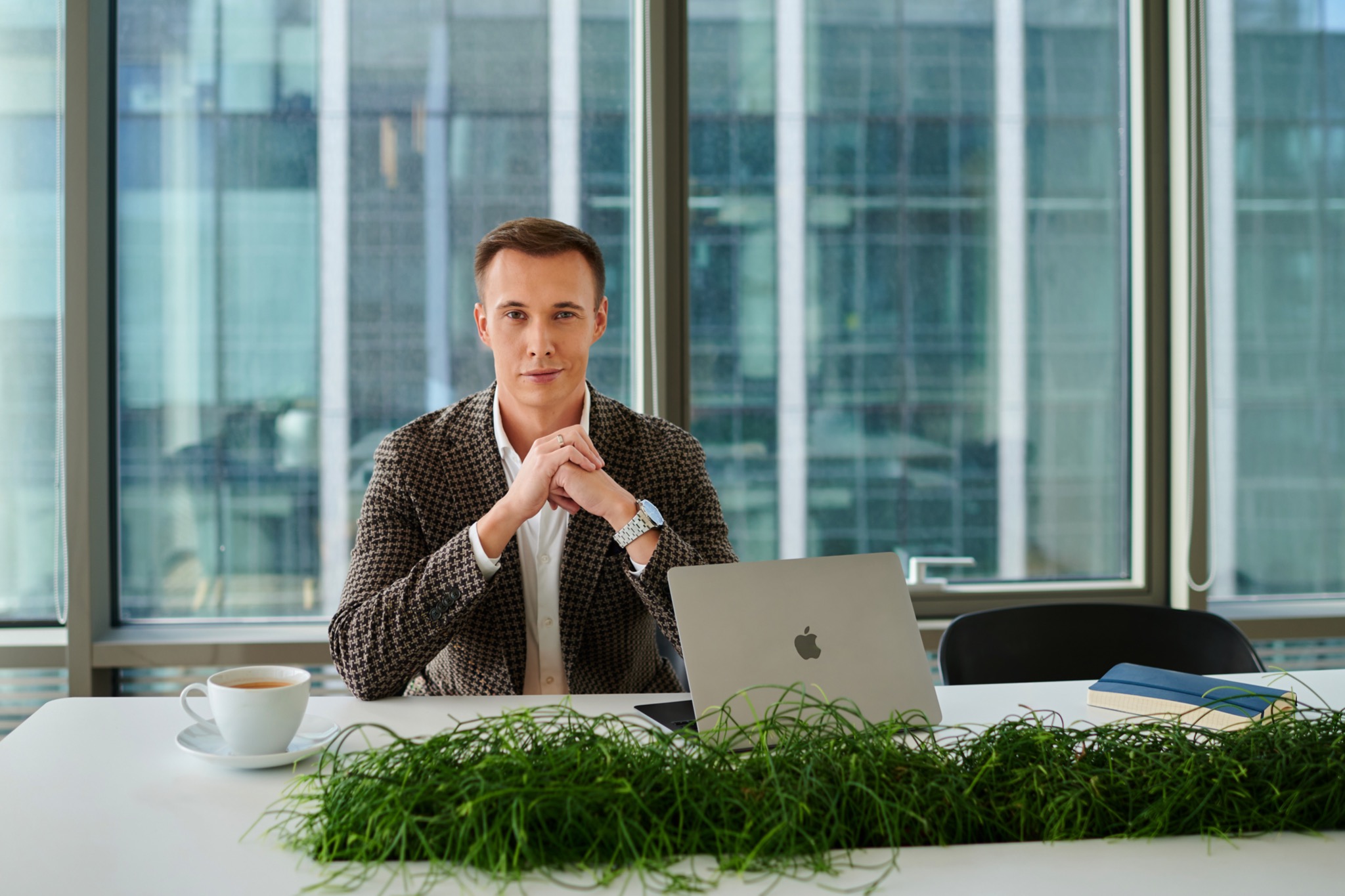 Młody mężczyzna w tweedowej marynarce siedzi za biurkiem z laptopem, kawą i rośliną, na tle przeszklonego biurowca.