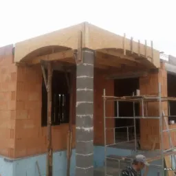 Wznoszenie ścian zewnętrznych domu z czerwonej cegły, widoczne drewniane szalunki nad oknami i filar z szarych pustaków, fragment rusztowania i dwóch robotników w kadrze.