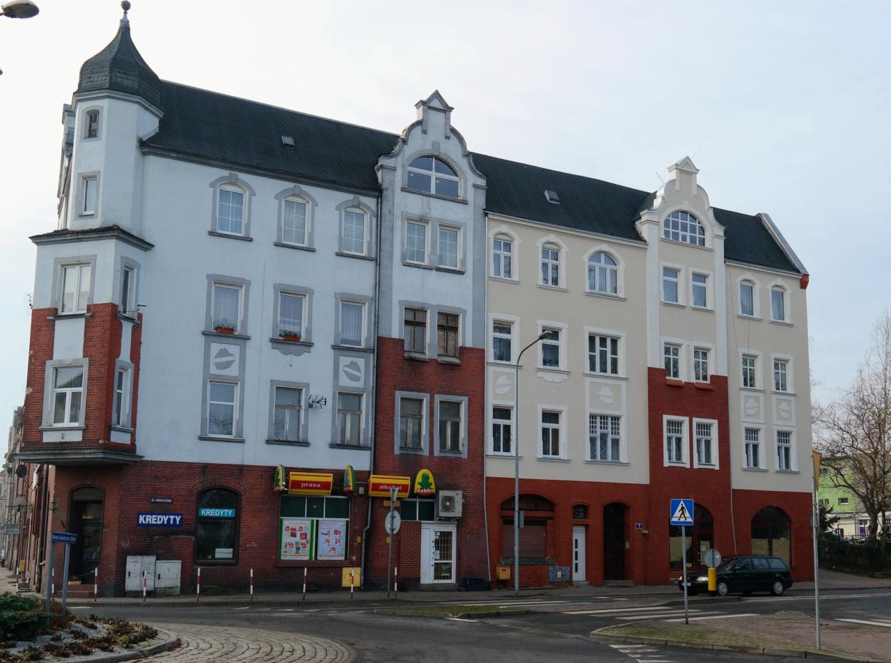 Fasada kamienicy z elementami muru pruskiego, pomalowana w pastelowych kolorach z czerwonymi akcentami na parterze, widok z ulicy na skrzyżowaniu z zaparkowanym czarnym samochodem.