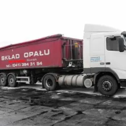 Ciężarówka z czerwoną naczepą z napisem 'SKŁAD OPAŁU' zaparkowana na terenie składu, widoczny śnieg i hałda węgla w tle.