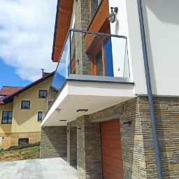 Spektrum technika - Nowoczesna balustrada ze szkła hartowanego na balkonie budynku obłożonego kamieniem elewacyjnym, widok z dołu na tle błękitnego nieba.