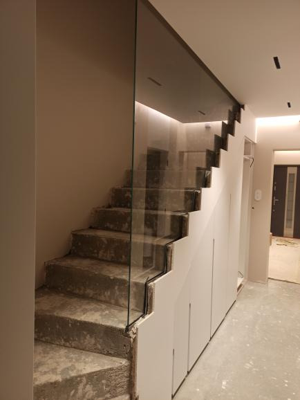 Szklana balustrada zamontowana na betonowych schodach w trakcie remontu, widoczne zabezpieczenia i niedokończone wykończenie.