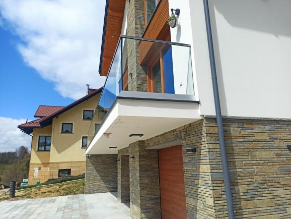 Nowoczesna balustrada ze szkła hartowanego na balkonie budynku obłożonego kamieniem elewacyjnym, widok z dołu na tle błękitnego nieba.