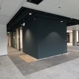 Wnętrze biurowe w trakcie wykańczania: pomalowana na ciemny kolor ściana, sufit z widocznymi instalacjami, betonowa podłoga z puszkami elektrycznymi, okna z grzejnikami.