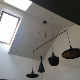 Wnętrze z białymi ścianami, skośnym sufitem z oknem dachowym i nowoczesną, czarną lampą wiszącą z czterema różnymi kloszami z elementami w kolorze złotym.