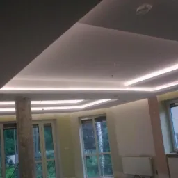 Kaskadowy sufit podwieszany z oświetleniem LED w jasnym pomieszczeniu z kolumną i dużymi oknami wychodzącymi na zieleń.