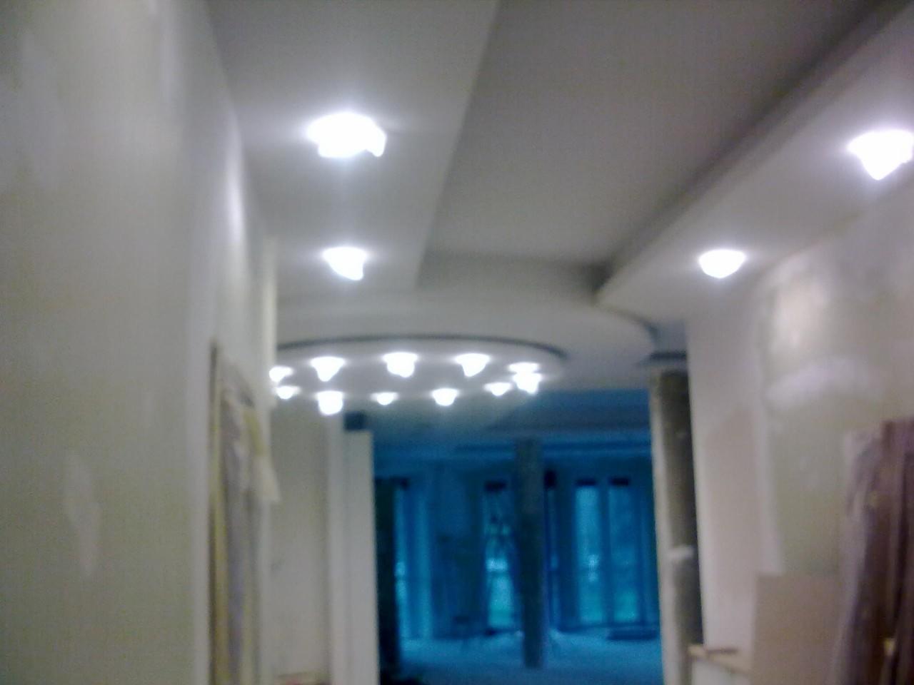 Korytarz w trakcie prac wykończeniowych, widoczne przygotowane ściany, sufit podwieszany z okrągłym wzorem i liczne wbudowane punkty świetlne, w tle niebieskie drzwi balkonowe.
