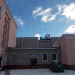 Budynek w pastelowych kolorach z widocznymi elementami wentylacyjnymi na dachu, zimowy krajobraz z resztkami śniegu na dziedzińcu i zaparkowanym zielonym samochodem.