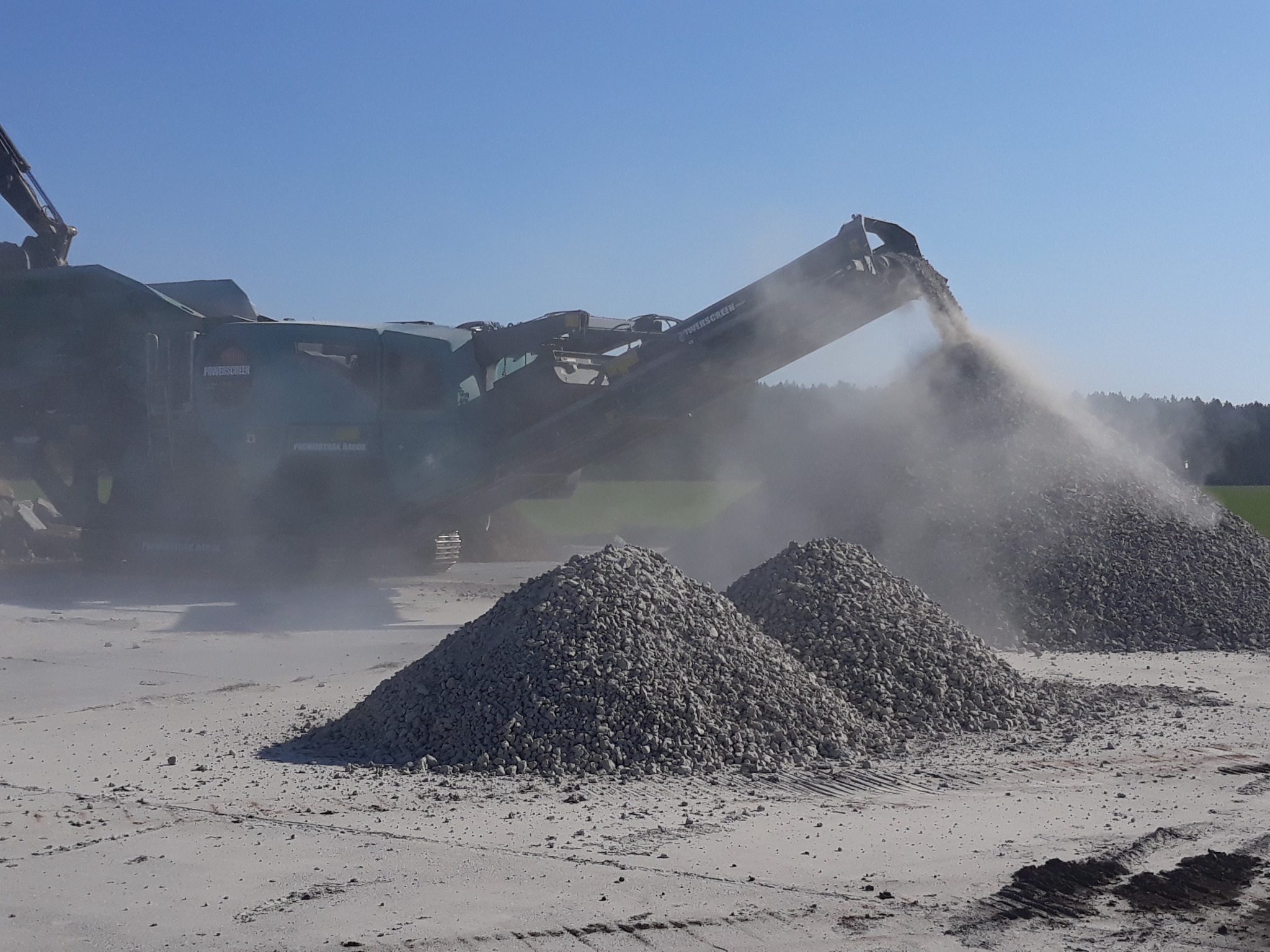 Praca kruszarki Powerscreen: hałdy gruzu na placu budowy, unoszący się pył i błękitne niebo w tle.