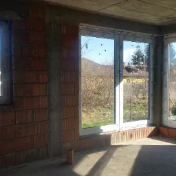 Wnętrze w stanie surowym z trzema oknami PCV marki Veka, widok na zabudowę jednorodzinną i drzewa za oknem.