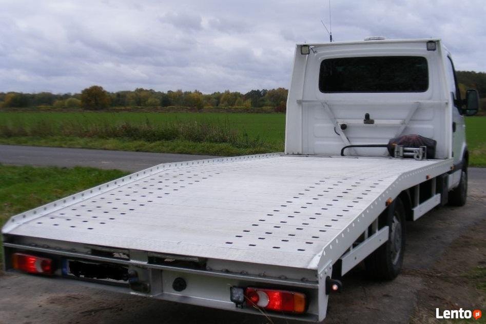 Widok z tyłu białej lawety z perforowaną platformą, na tle zielonego pola i pochmurnego nieba.