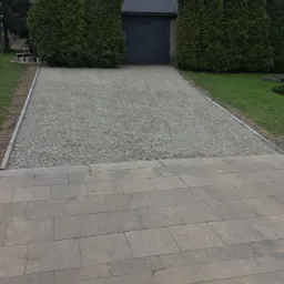 Szeroki podjazd z jasnego żwiru, obrzeża z szarej kostki, przejście do chodnika z regularnych płyt, w tle budynek gospodarczy i wysokie tuje.