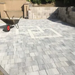 Ułożona szara kostka brukowa z dekoracyjnym wzorem jasnej kostki w Grodzisku, widoczna taczka i szpadel przy murku oporowym.