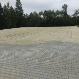 Anna Krawczyk P.U.H. Bartotex - Szara kostka brukowa ułożona na dużej powierzchni, z przygotowanym podłożem w tle i zielenią drzew. Widok z góry, pochmurne niebo.