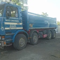 Niebieska ciężarówka Scania z wywrotką, z widocznym logo firmy Arved Christensen, zaparkowana na placu budowy, w tle biały MAN i koparka.