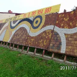 Ekspozycja wzorów nawierzchni z kostki brukowej i granitowej, prezentacja różnorodnych kolorów i układów, na tle zielonej trawy i budynku. Baner reklamowy z numerem telefonu widoczny w górnej...