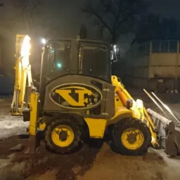 Żółto-szara koparko-ładowarka z logo firmy na tle nocnego, oświetlonego placu budowy z zaparkowanymi pojazdami.