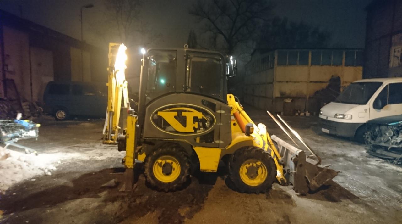 Żółto-szara koparko-ładowarka z logo firmy na tle nocnego, oświetlonego placu budowy z zaparkowanymi pojazdami.