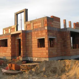 Budowa domu jednorodzinnego z czerwonej cegły, widoczne otwory okienne i drzwiowe, częściowo wzniesione ściany, palety z cegłami na placu budowy.