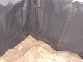 Wnętrze wykopu wyłożonego czarną folią, wypełnionego częściowo piaskiem i ziemią. Kąt ujęcia z góry, ukazujący strukturę podłoża i zabezpieczenie folią.