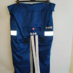 Niebieskie spodnie robocze na wieszaku z odblaskowymi paskami i symbolami na kieszeniach.