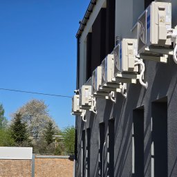 Termi - Siedem zewnętrznych jednostek klimatyzacji Haier, zamontowanych w rzędzie na ciemnoszarej, teksturowanej elewacji budynku. Widoczne białe izolowane rury i długie cienie w słoneczny dzień.