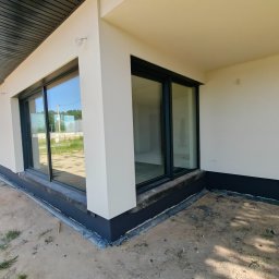 Termi - Narożnik nowo wybudowanego domu z jasnym tynkiem, dużymi, ciemnymi oknami przesuwnymi i widoczną czarną izolacją fundamentów. Ciemna podbitka dachu z rynną.