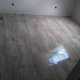 Mar Bud - Wyłożona płytkami podłoga w łazience, imitująca drewno, z widocznymi przyłączami do baterii na ścianie i odbiciem światła na powierzchni