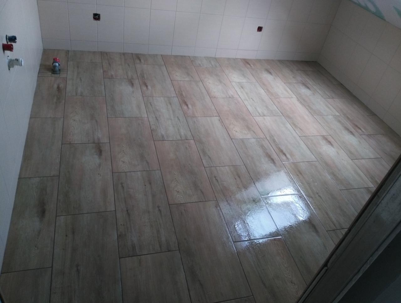 Wyłożona płytkami podłoga w łazience, imitująca drewno, z widocznymi przyłączami do baterii na ścianie i odbiciem światła na powierzchni