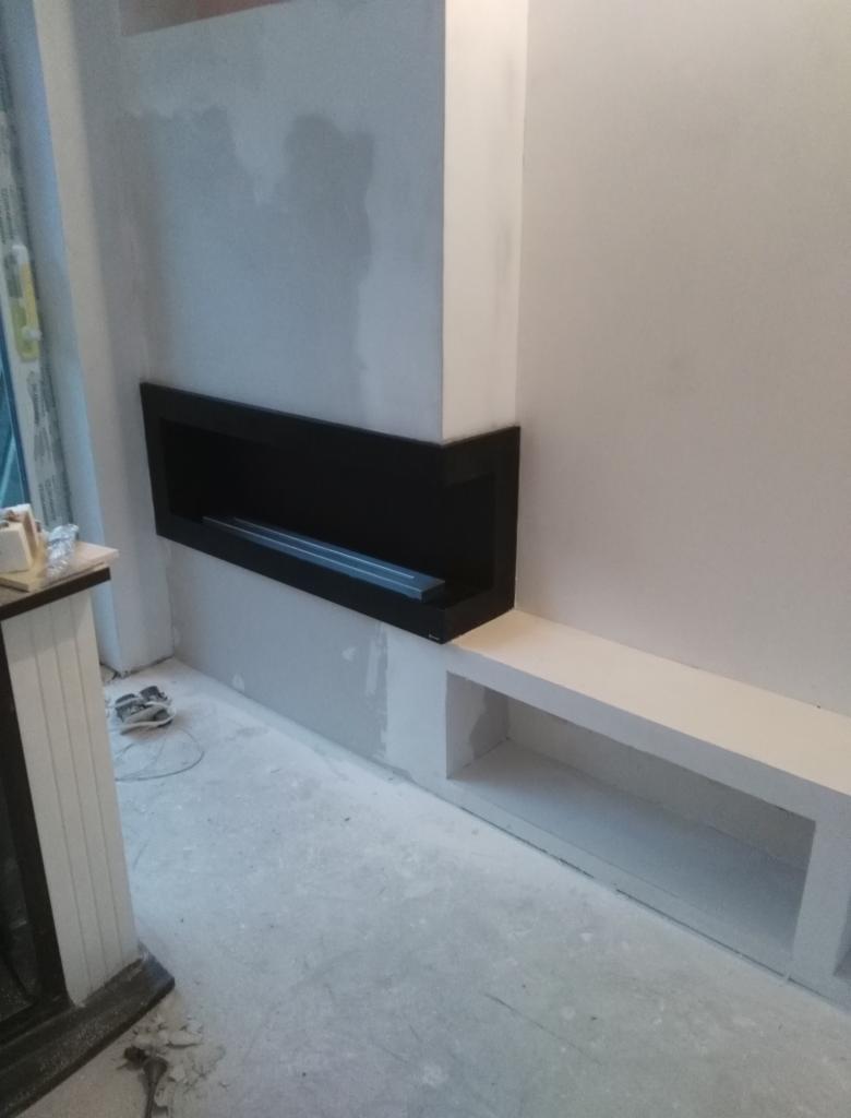 Ściana w trakcie wykańczania z wnęką na kominek w czarnej obudowie i białą wnęką obok, surowy beton na podłodze, widoczne ślady po szpachlowaniu na ścianach.
