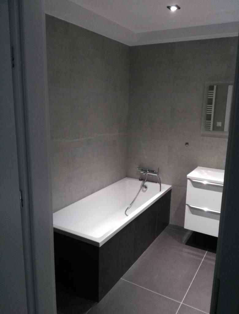 Nowoczesna łazienka z wanną obudowaną ciemnymi płytkami, szare płytki ścienne i podłogowe, biała umywalka z szafką i grzejnik drabinkowy.