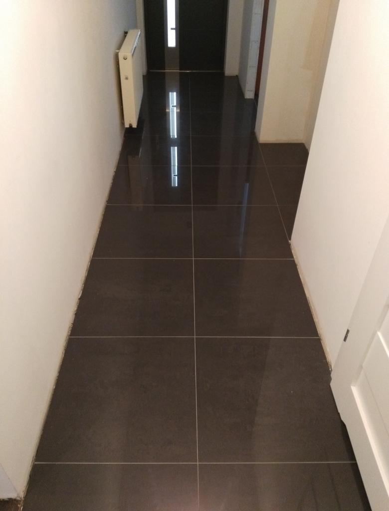 Położona ciemna, błyszcząca podłoga z płytek w wąskim korytarzu z białymi ścianami i grzejnikiem.