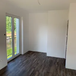 Pusty pokój z ciemnobrązowymi panelami podłogowymi, świeżo pomalowanymi na biało ścianami i balkonem z widokiem na zieleń.